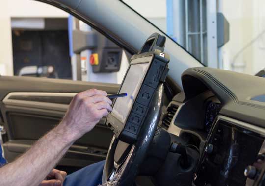 Un empleado de tienda de repuestos de automóviles revisando el inventario y utilizando una tablet para gestionar las existencias, parado al lado de estanterías llenas de cajas de repuestos.